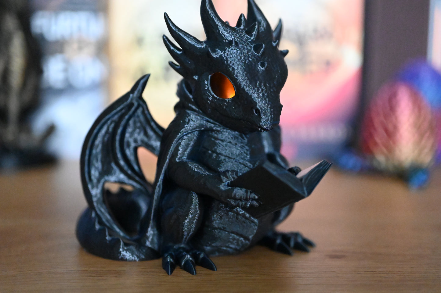 Figurina dragon negru cu ochi luminați portocaliu, citind o carte pe o masă din lemn, decor fantasy mistic pentru pasionații de cărți și povești cu dragoni.
