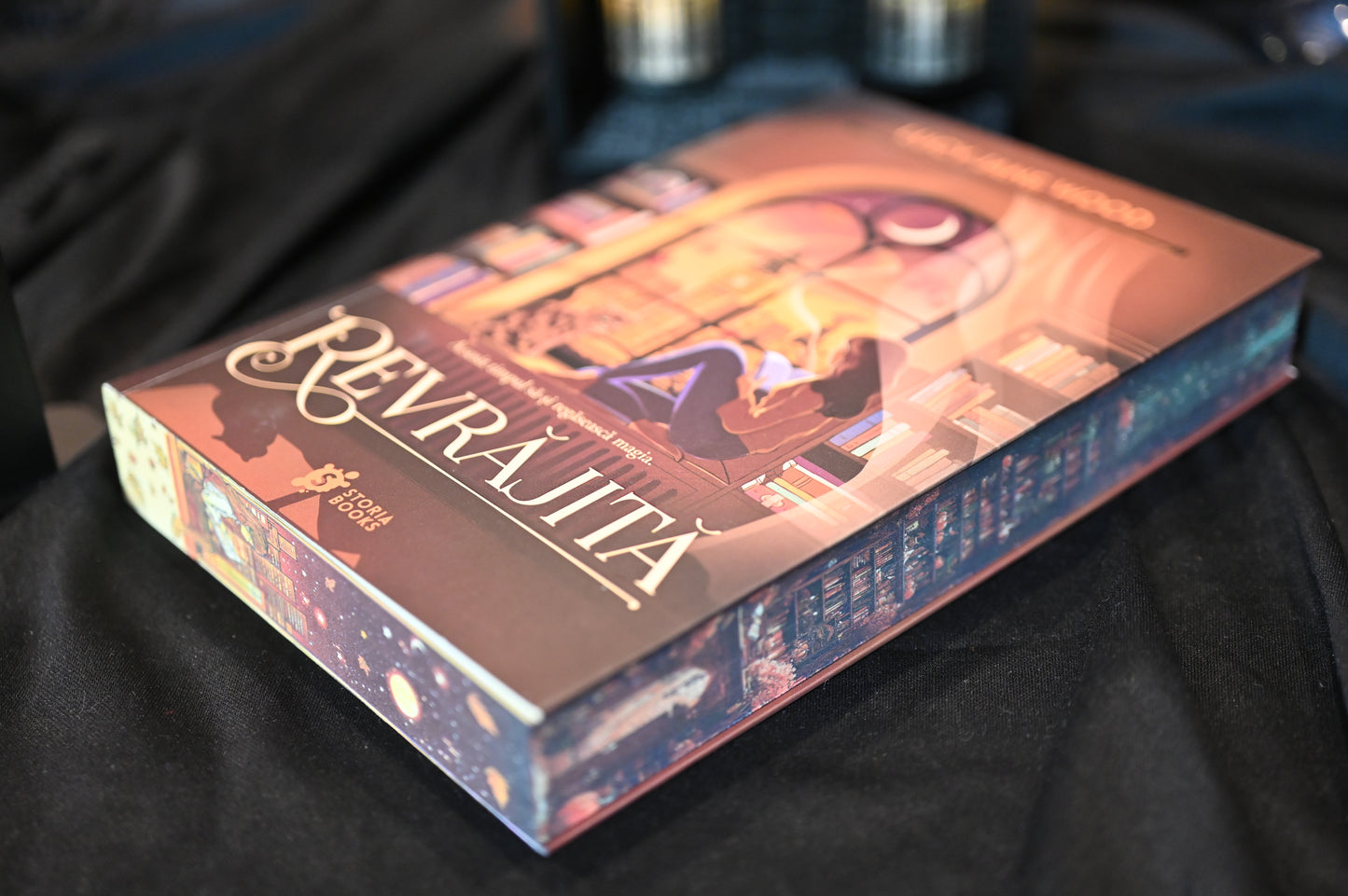 Cartea Revrăjita de Lucy Jane Wood, fotografiată în prim-plan pe un fundal întunecat. Coperta arată o tânără care citește lângă o fereastră luminată de lună, iar pe margini se văd detalii ilustrate cu rafturi de cărți și motive cosmice, în nuanțe de mov și cupru.