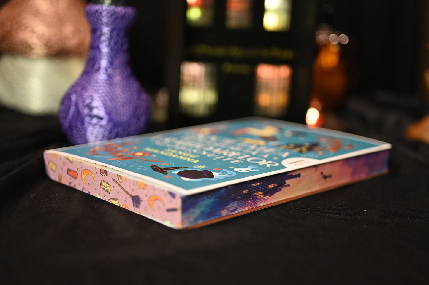 Cartea Societatea secretă a vrăjitoarelor de Sangu Mandanna, fotografiată în poziție orizontală. Coperta albastră vibrantă și marginea imprimată cu motive de magie, cărți și cer în nuanțe calde, accentuează tema fantasy și atmosfera cozy a ediției cu printed edges.