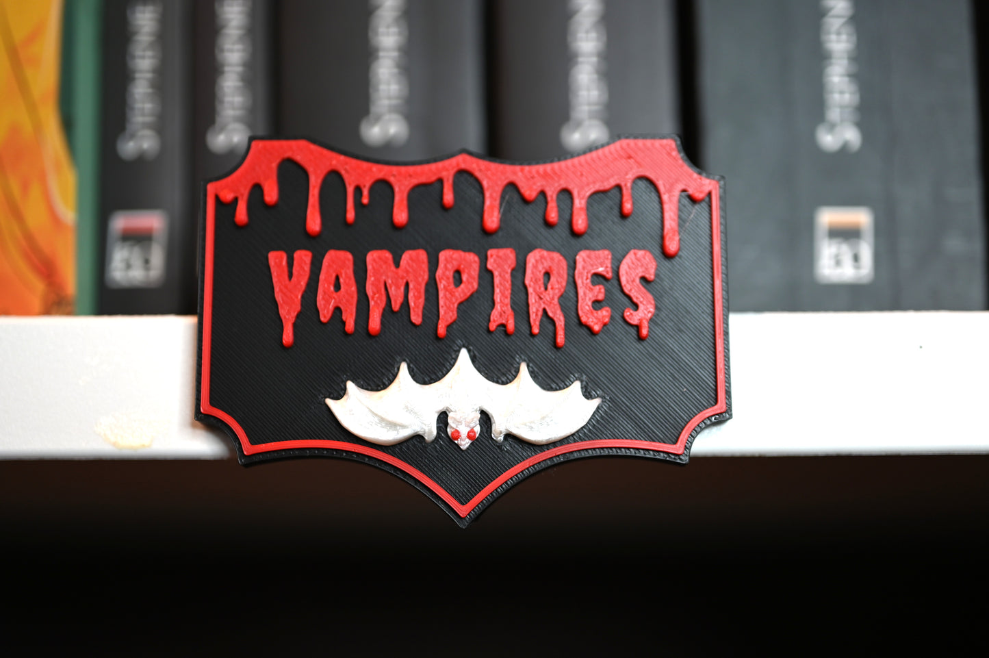 Etichetă 3D Vampires, neagră cu margini roșii, litere roșii care par scurse și un liliac alb cu ochi roșii în partea de jos