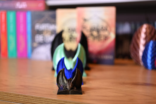 Decor dragon pentru bibliotecă- negru, verzui, albastru si auriu - cu aripi mari, stând pe o cate carte deschisă, iar in fundal sunt  cărțile din seria Rebecca Yarros, inclusiv ‘A Patra Aripă’ și ‘Flacăra de Fier’.