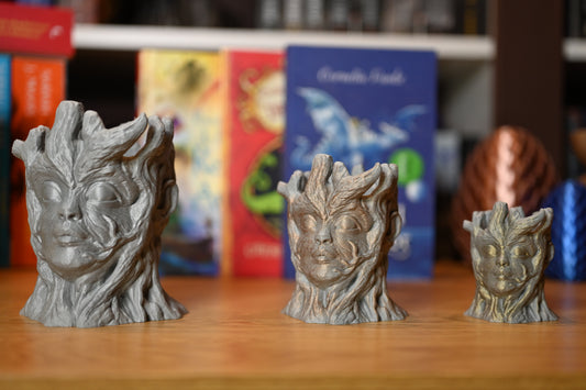 Trei suporturi decorative în formă de fețe sculptate cu trăsături de copac, realizate în stil fantasy, așezate pe o masă de lemn, cu cărți colorate pe fundal.