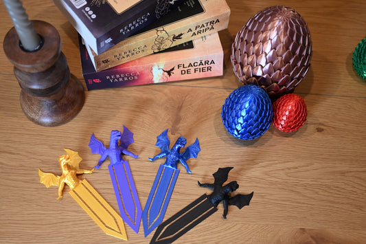 Semn de carte dragon 3D în formă de dragon, colorate în auriu, violet, albastru și negru, așezate pe o masă lângă ouă decorative de dragon și cărțile scrise de Rebecca Yarros