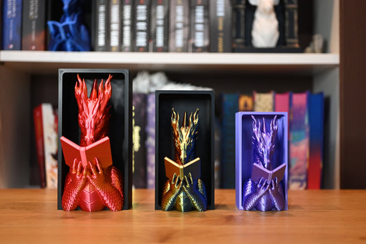 Trei suporturi fantasy de cărți în formă de dragoni care citesc, în culori roșu, auriu-verzui și mov, așezate pe un raft de lemn, cu fundal de bibliotecă fantasy.