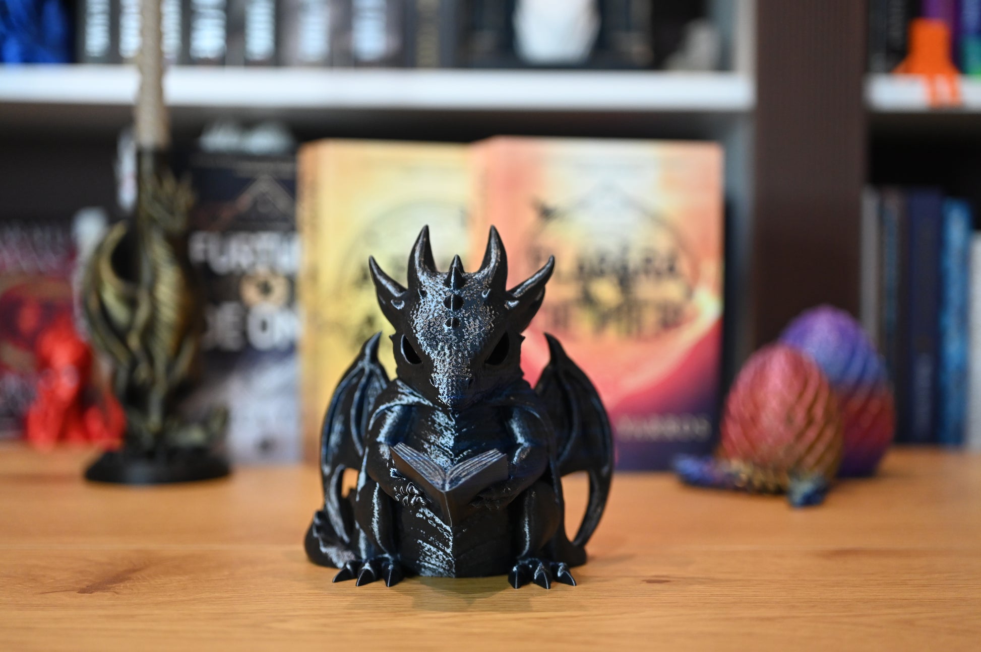 Dragonul Cititor Figurina dragon albastru cu detalii metalice, ținând o carte, decor fantasy pentru bibliotecă sau raft de cărți, pe fundal cu cartile scrise de Rebecca Yarros