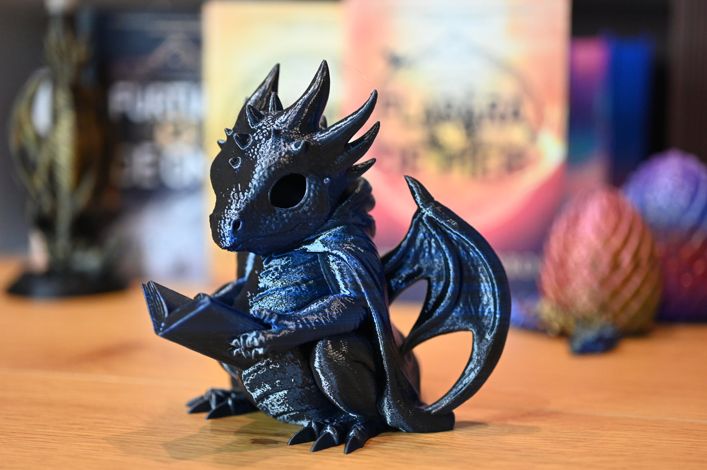 Dragonul Cititor Figurina dragon albastru cu detalii metalice, ținând o carte, decor fantasy pentru bibliotecă sau raft de cărți, pe fundal cu cartile seriei A patra aripa