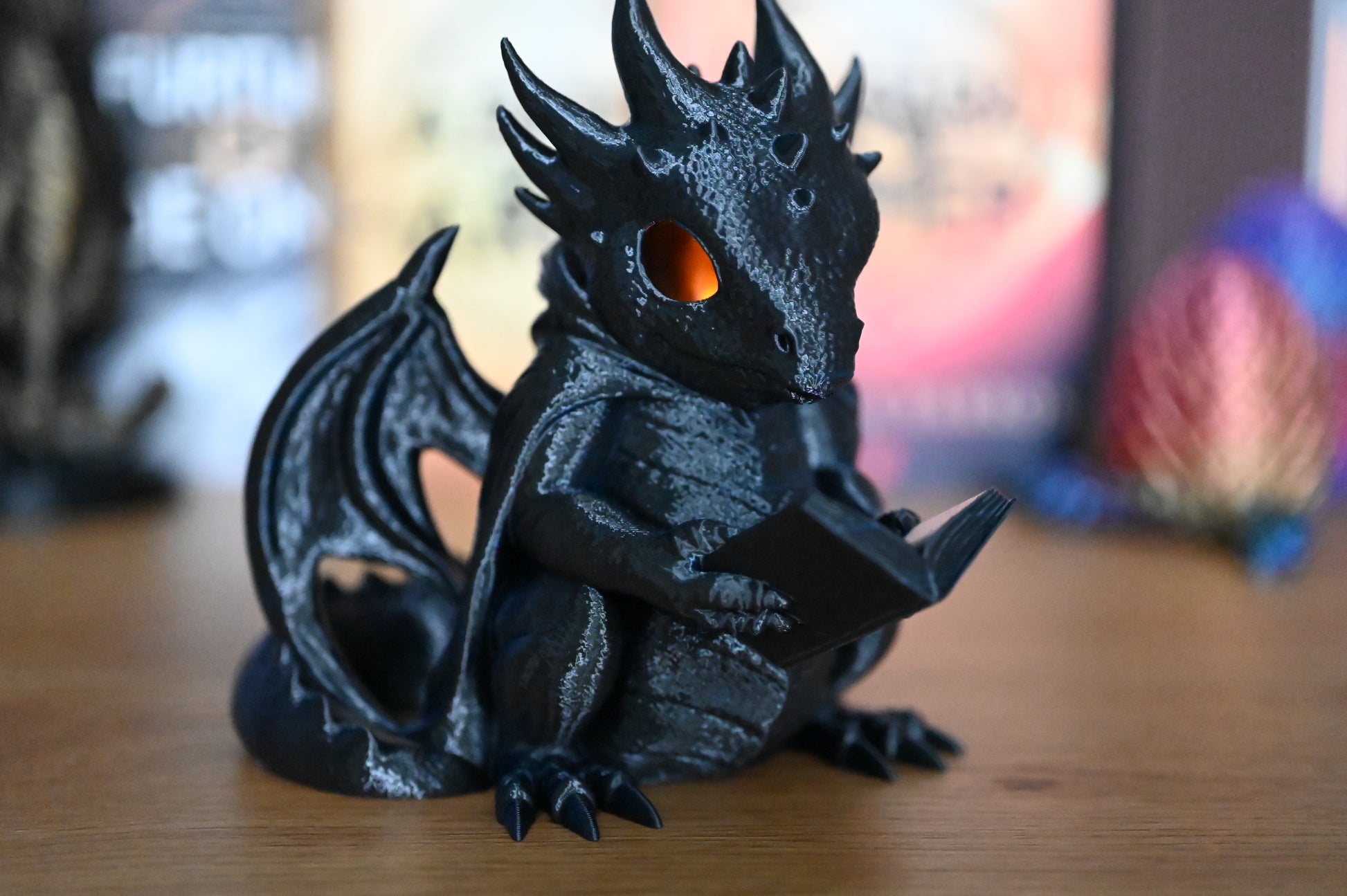 Figurina dragon negru cu ochi luminați portocaliu, citind o carte pe o masă din lemn, decor fantasy mistic pentru pasionații de cărți și povești cu dragoni.