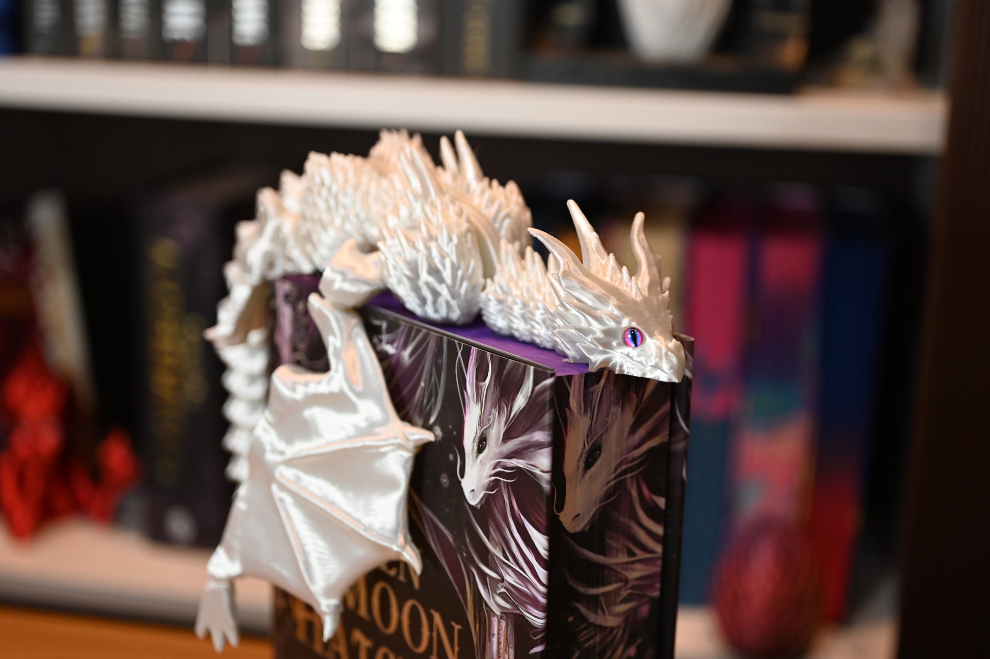 Dragon alb strălucitor cu ochi violet, așezat pe o carte cu copertă ilustrată, decor fantasy pentru iubitorii de lectură magică.
