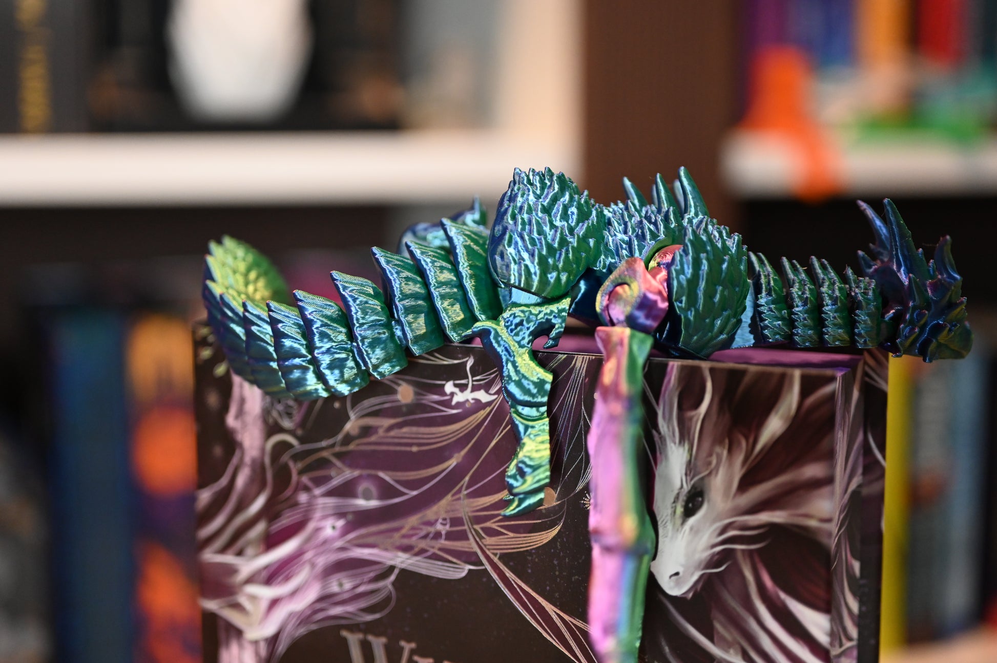Figurina dragon iridescent în nuanțe de verde și albastru, cu aripi deschise, așezat pe o carte, decor de inspirație fantasy.