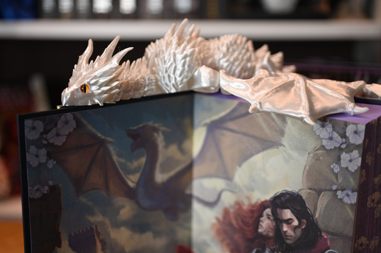 Dragon alb sidefat cu ochi portocalii, odihnindu-se pe o carte ilustrată cu dragoni, decor fantasy de inspirație epică.