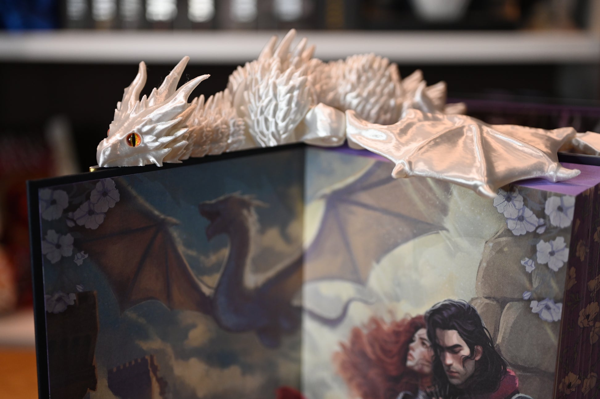 Dragon alb sidefat cu ochi portocalii, odihnindu-se pe o carte ilustrată cu dragoni, decor fantasy de inspirație epică.