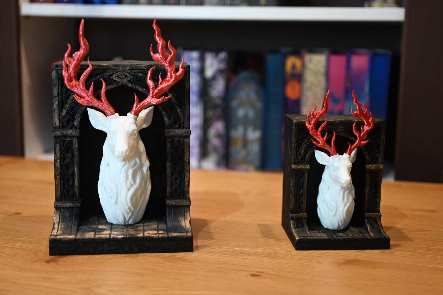 Două suporturi de cărți în stil fantasy, reprezentând busturi de cerb alb cu coarne roșii în formă de flăcări, montate pe baze negre sculptate, așezate pe o masă din lemn.