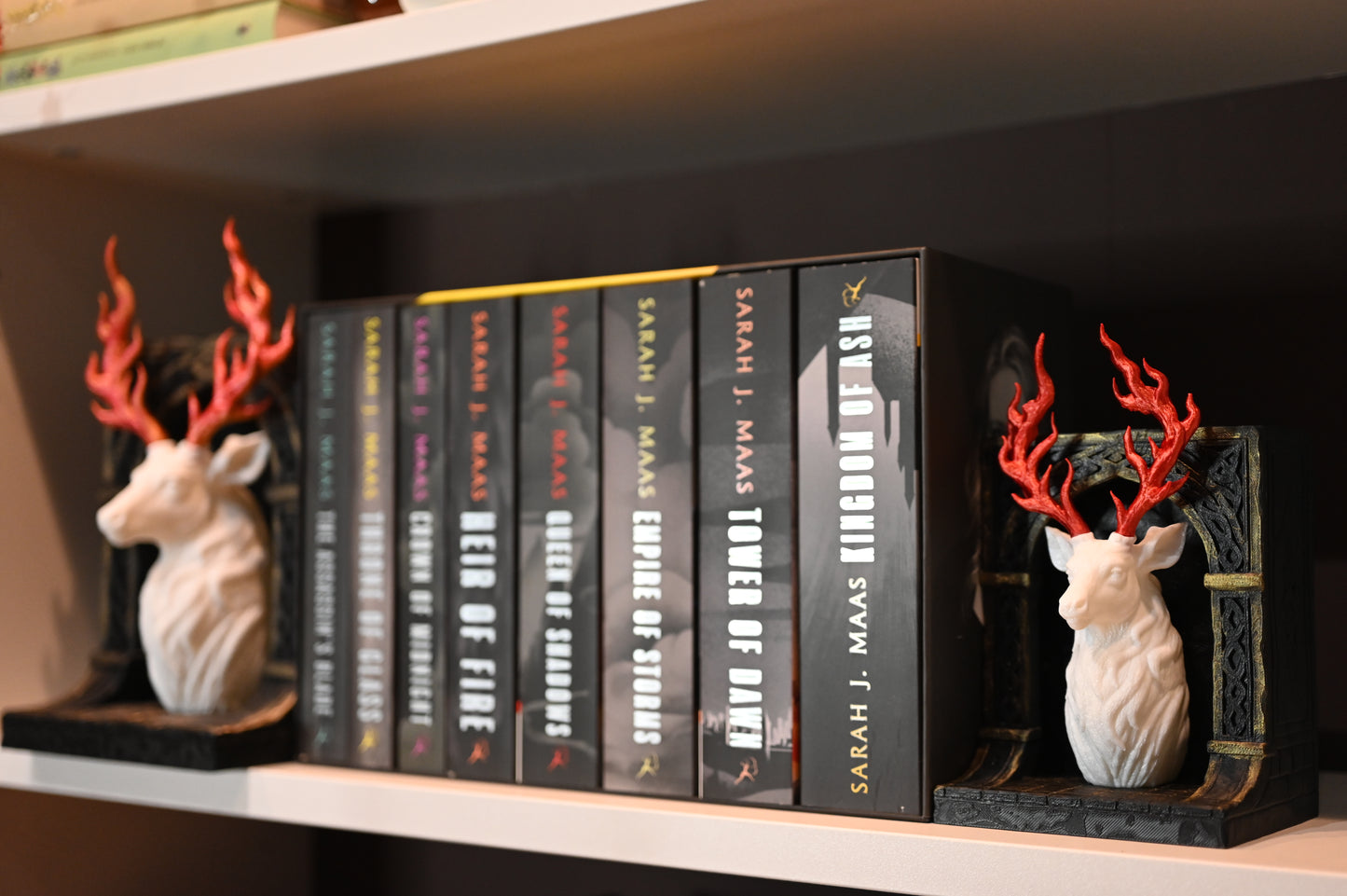 Două suporturi de cărți în stil fantasy, reprezentând busturi de cerb alb cu coarne roșii în formă de flăcări, montate pe baze negre sculptate, așezatein biblioteca, langa cartile seriei Tronul de clestar