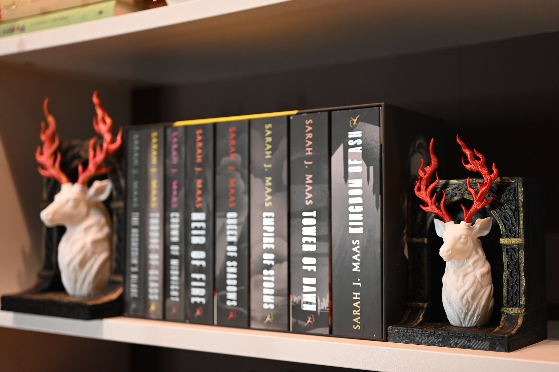 Două suporturi de cărți în stil fantasy, reprezentând busturi de cerb alb cu coarne roșii în formă de flăcări, montate pe baze negre sculptate, așezatein biblioteca, langa cartile seriei Tronul de clestar