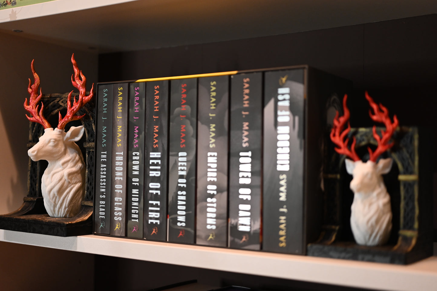 Două suporturi de cărți în stil fantasy, reprezentând busturi de cerb alb cu coarne roșii în formă de flăcări, montate pe baze negre sculptate, așezate pe un raft cu cartile scrise de Sarah J maas, seria Tronul de clestar