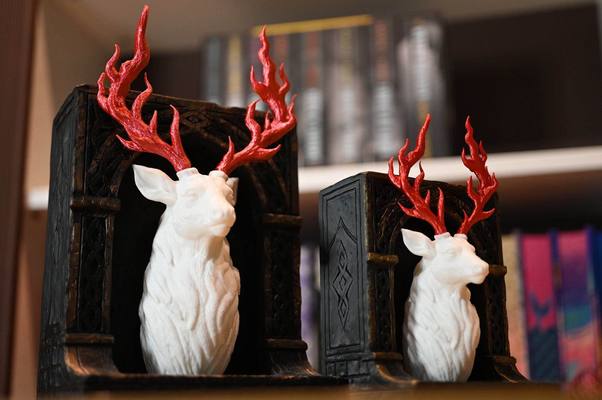 Două suporturi de cărți în stil fantasy, reprezentând busturi de cerb alb cu coarne roșii în formă de flăcări, montate pe baze negre sculptate, așezate pe o masă din lemn.