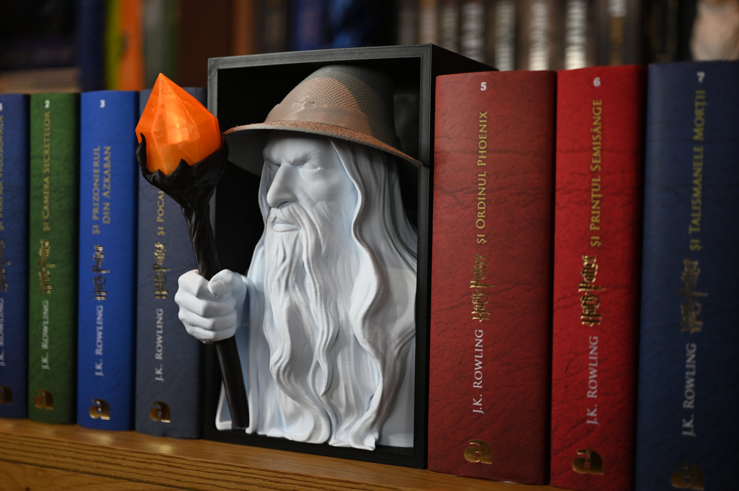 Vrăjitorul Luminii - Book nook Bust 3D al unui vrăjitor inspirat de Gandalf The Grey, cu pălărie și barbă lungă, ținând un toiag cu cristal portocaliu, plasat între volumele colorate ale seriei Harry Potter pe un raft de cărți.