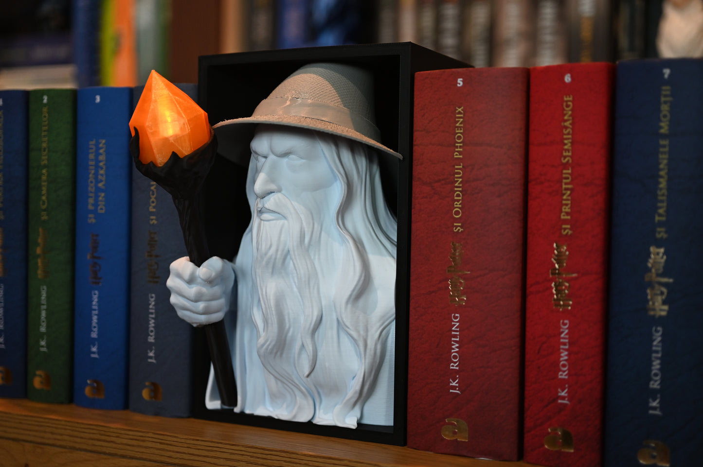 Vrăjitorul Luminii - Book nook Bust 3D al unui vrăjitor cu pălărie și barbă lungă, ținând un toiag cu cristal portocaliu, plasat între volumele colorate ale seriei Harry Potter pe un raft de cărți.