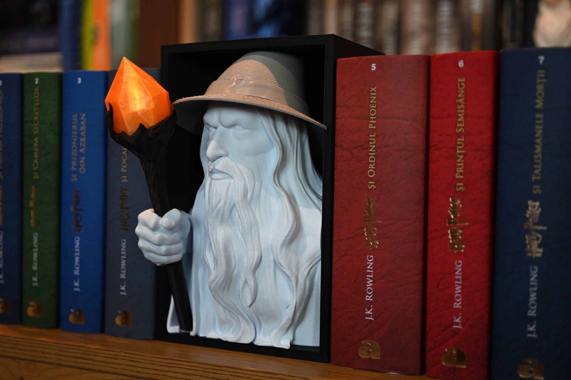 Vrăjitorul Luminii - Book nook Bust 3D al unui vrăjitor cu pălărie și barbă lungă, ținând un toiag cu cristal portocaliu, plasat între volumele colorate ale seriei Harry Potter pe un raft de cărți.