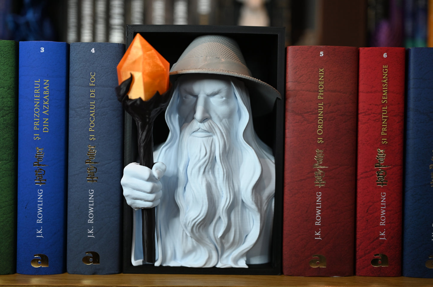 Vrăjitorul Luminii - Book nook Bust 3D al unui vrăjitor cu pălărie și barbă lungă, ținând un toiag cu cristal portocaliu, plasat între volumele colorate ale seriei Harry Potter pe un raft de cărți.