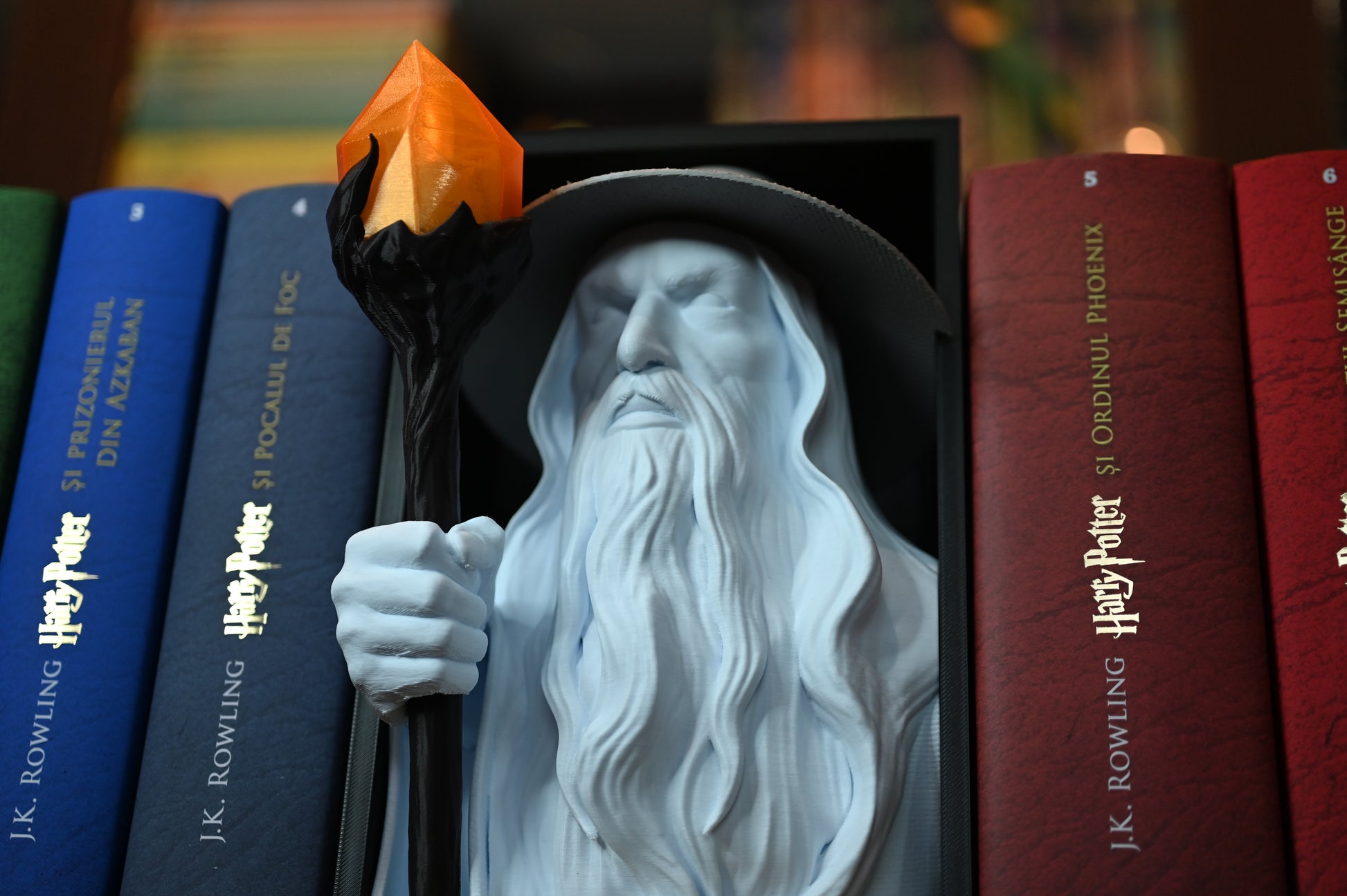 Bust 3D al unui vrăjitor inspirat din universul Hogwarts, cu pălărie și barbă lungă, ținând un toiag cu cristal portocaliu, plasat între volumele colorate ale seriei Harry Potter pe un raft de cărți.