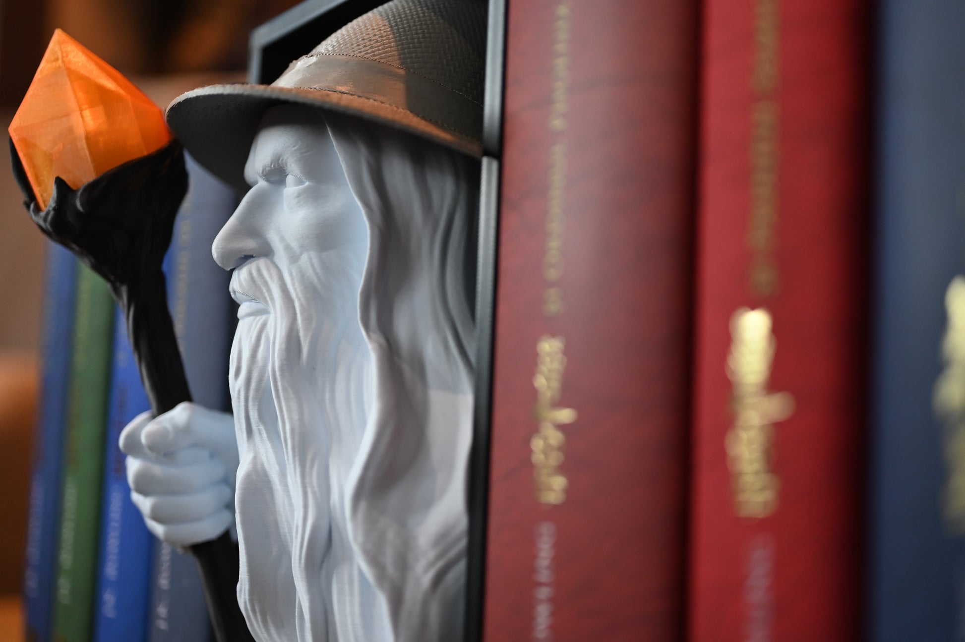 Bust 3D al unui vrăjitor cu pălărie și barbă lungă, ținând un toiag cu cristal portocaliu, plasat între volumele colorate ale seriei Harry Potter pe un raft de cărți.