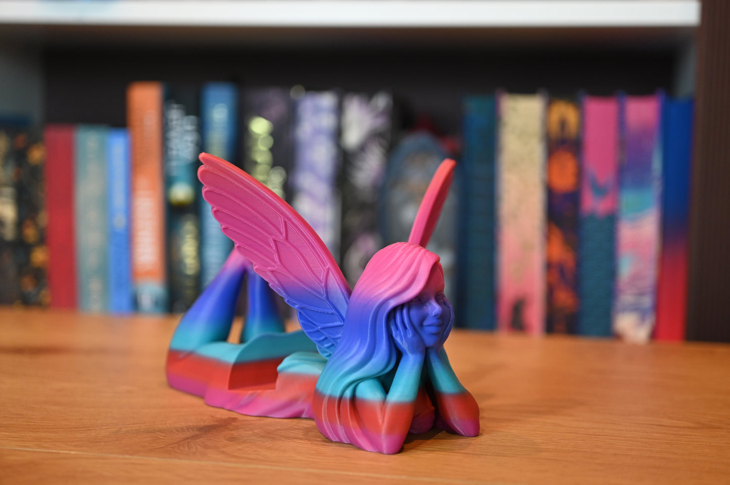 Suport pentru Kindle sau cărți - Zână în nuanțe de albastru, mov și roz, reprezentând o zână cu aripi mari roz, întinsă pe burtă și sprijinită în palme, pe un fundal de raft cu cărți fantasy.