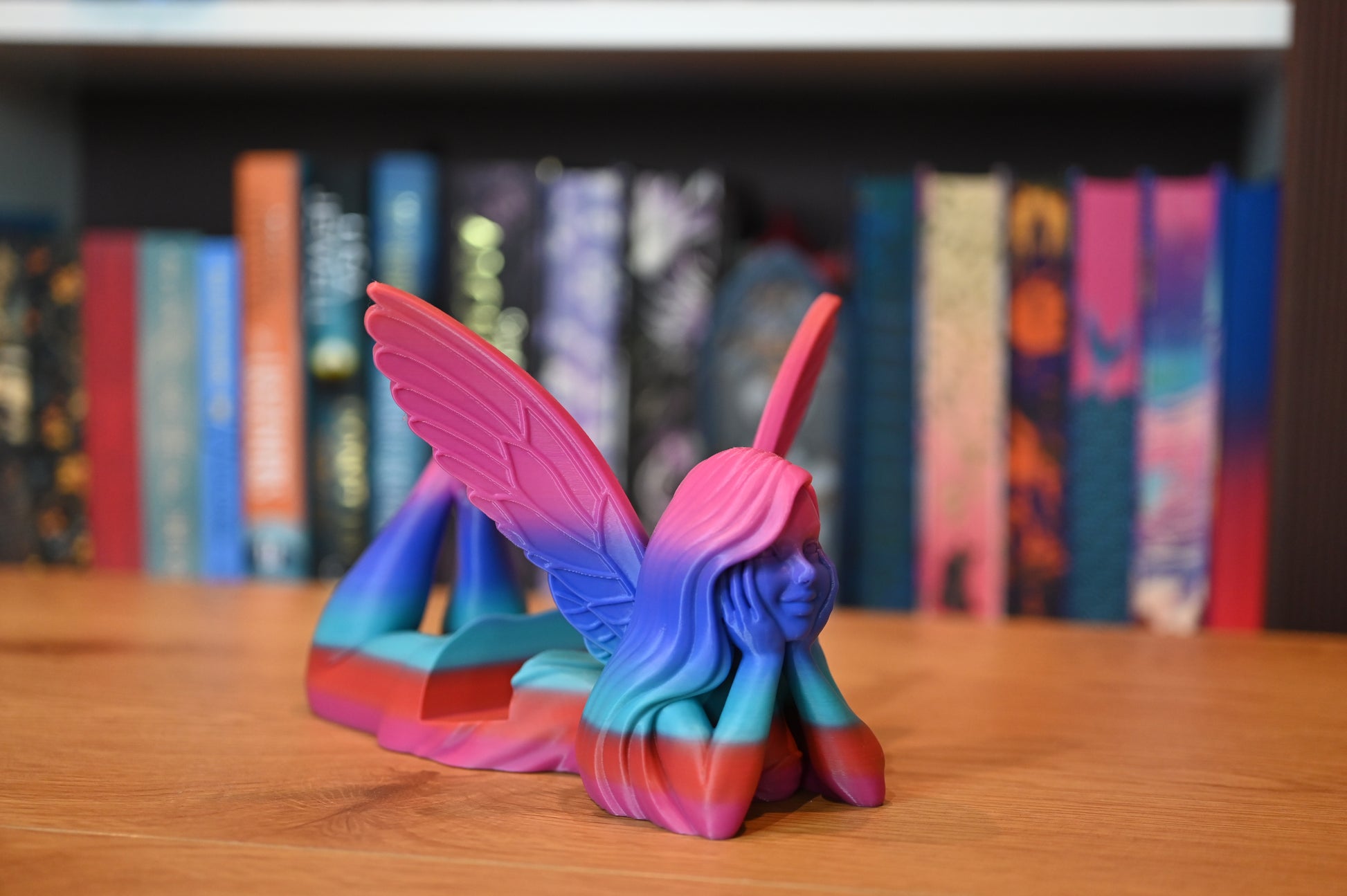 Suport pentru Kindle sau cărți - Zână în nuanțe de albastru, mov și roz, reprezentând o zână cu aripi mari roz, întinsă pe burtă și sprijinită în palme, pe un fundal de raft cu cărți fantasy.