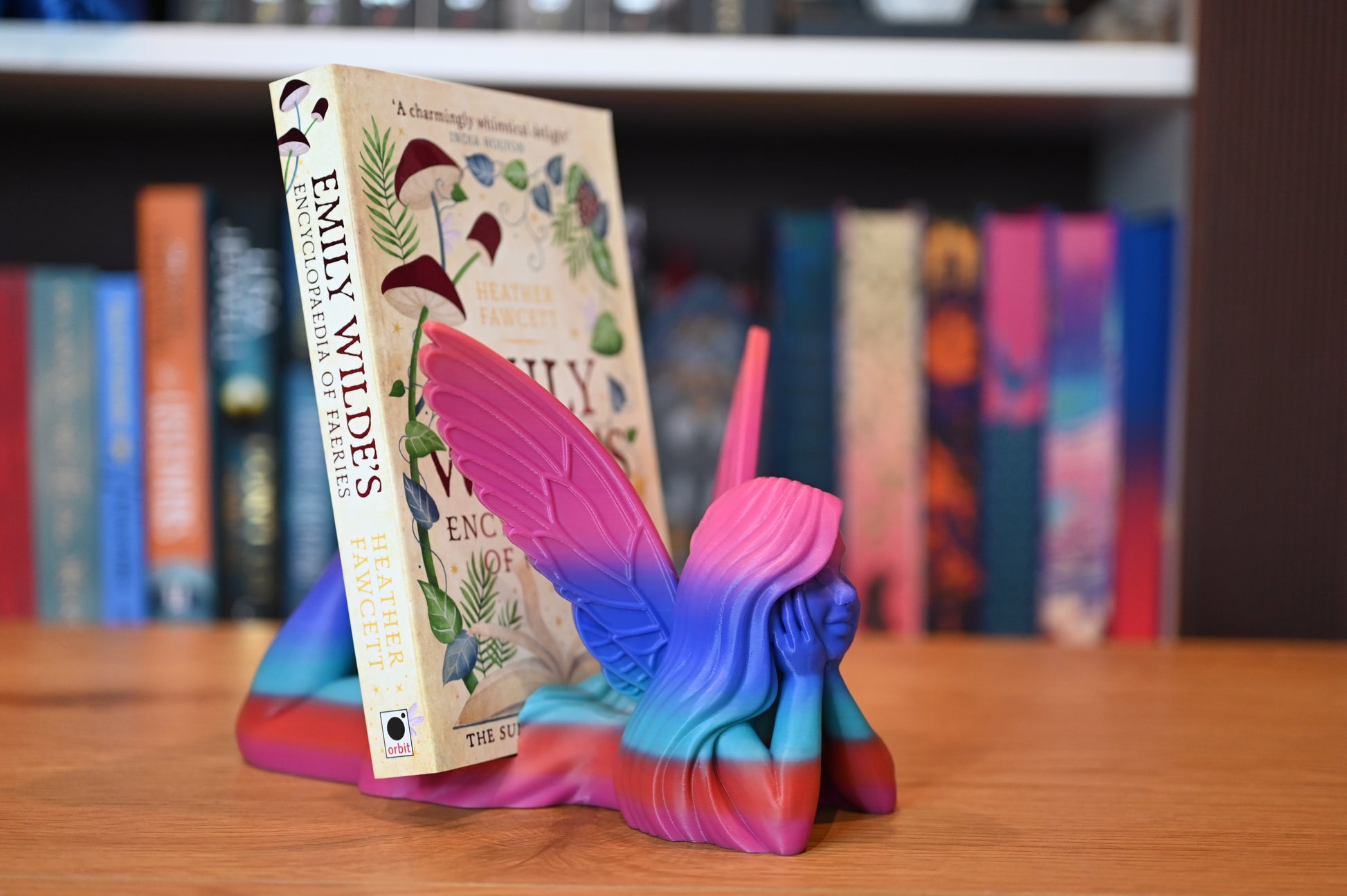 Suport pentru Kindle sau cărți - Zână multicoloră, cu aripi roz și corp în degrade albastru-mov-roșu, ținând o carte fantasy Enciclopedia Spiridusilor între aripi, pe un raft din lemn.