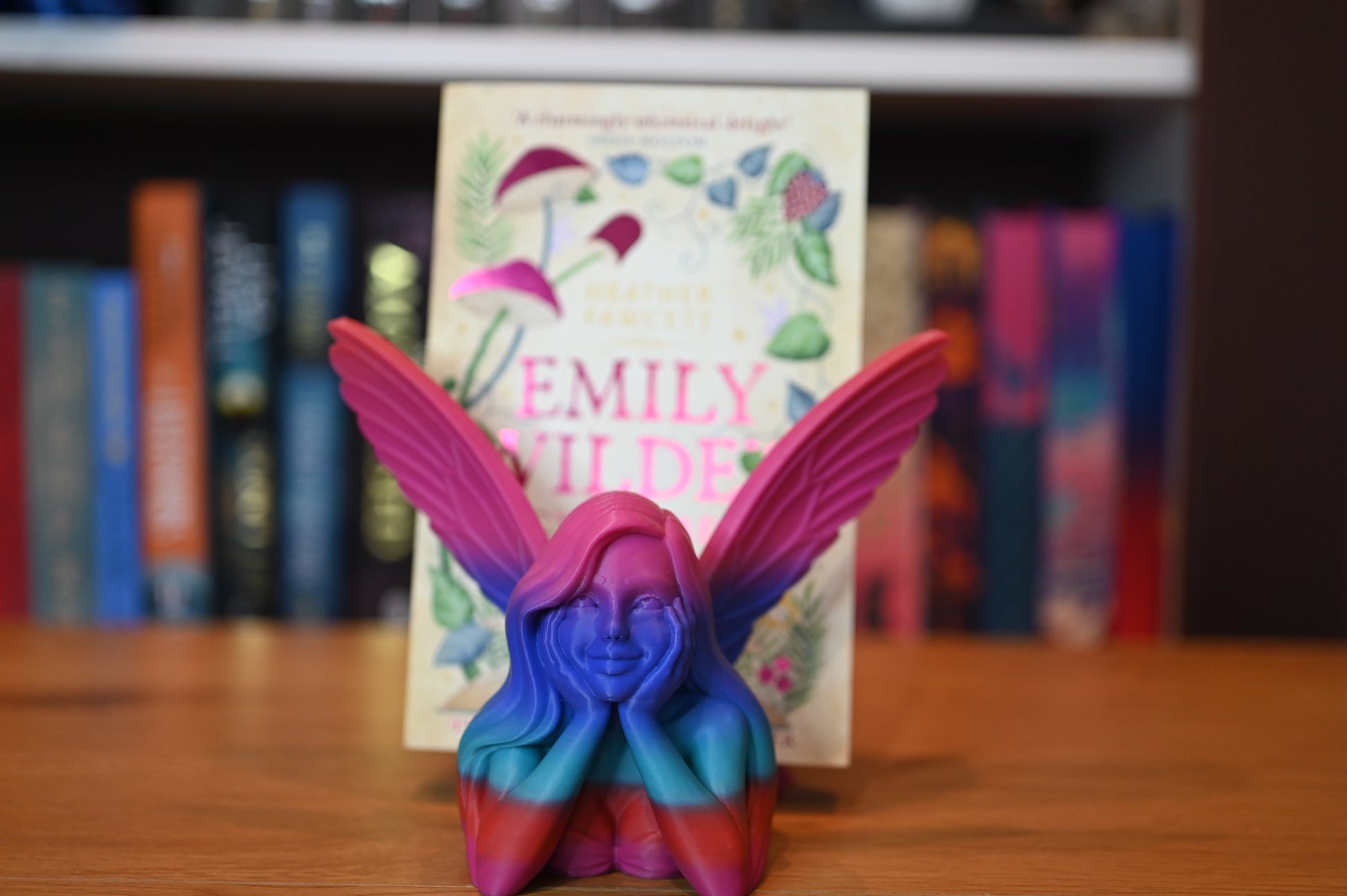 Vedere frontală a unei zâne în culori vibrante (albastru, roz, roșu), cu aripi largi deschise și zâmbet calm, plasată în fața cărții „Emily Wilde’s Encyclopaedia of Faeries”.