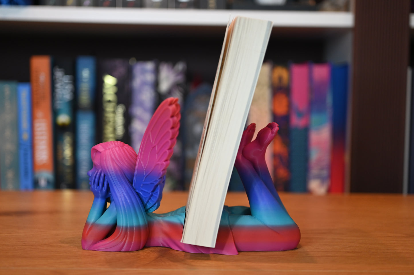 Vedere laterala a unei zâne în culori vibrante (albastru, roz, roșu), cu aripi largi deschise și zâmbet calm, plasată în fața cărții „Emily Wilde’s Encyclopaedia of Faeries”.