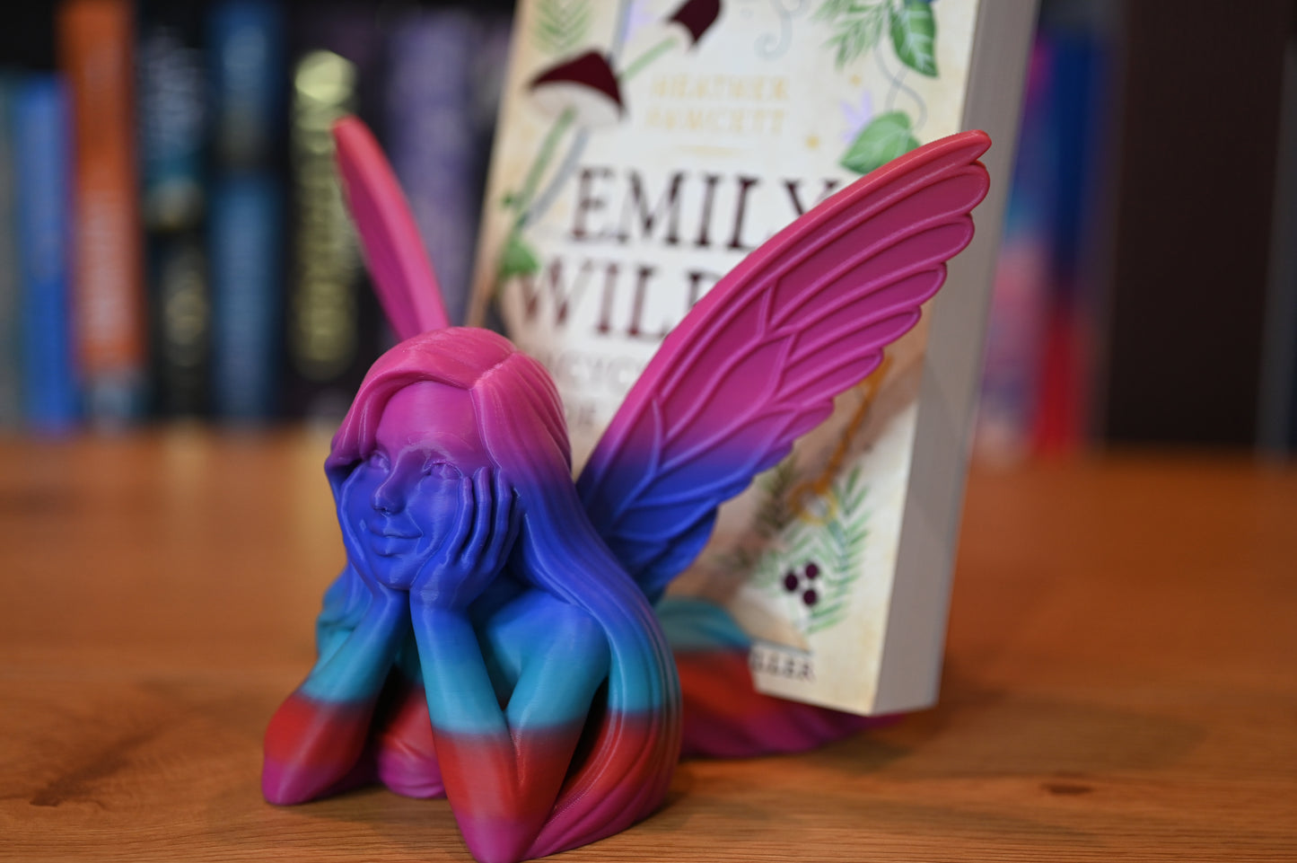 Suport de carte în formă de zână multicoloră, cu aripi roz și corp în degrade albastru-mov-roșu, ținând o carte fantasy între aripi, pe un raft din lemn.