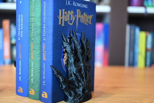 Suport de Cărți - Mână scheleticăîn formă de mână întunecată, cu aspect gotic, care ține în poziție trei volume Harry Potter, pe un fundal de bibliotecă.