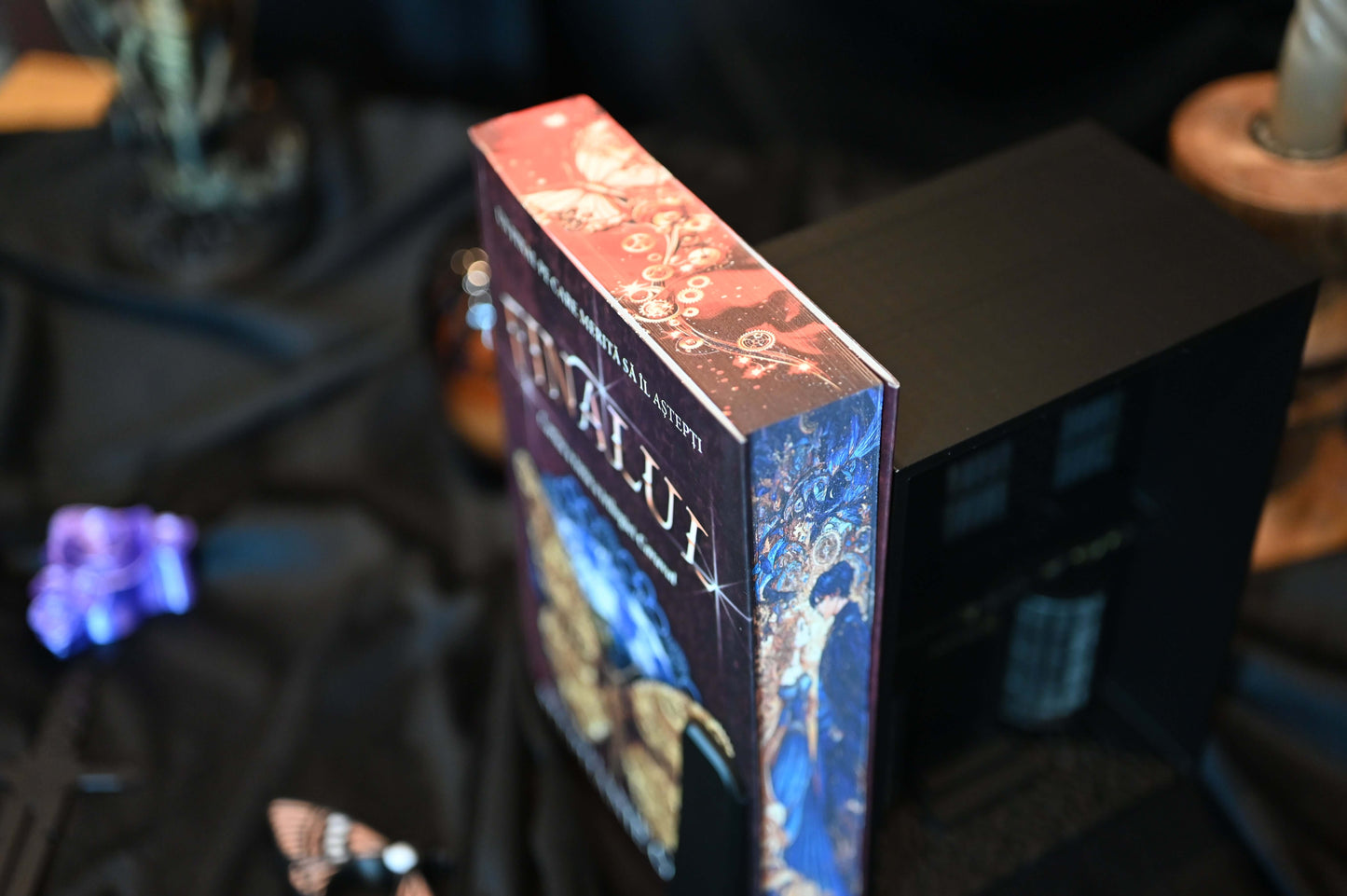 Vedere de sus a cărții Finalul, arătând designul imprimat pe muchii cu fluturi și rotițe aurii, în nuanțe de roșu și auriu.