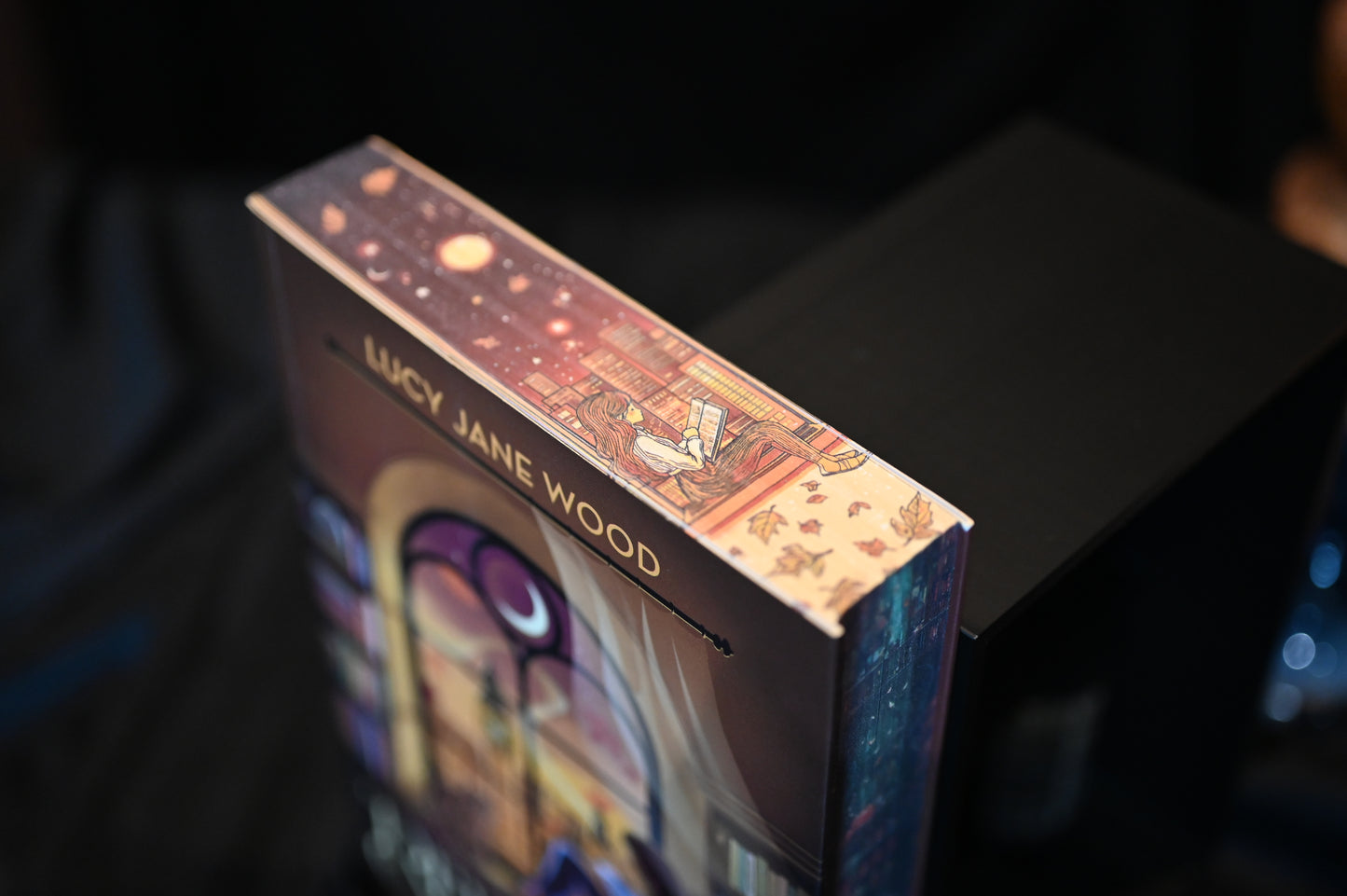 Vedere de sus a cărții Revrăjita de Lucy Jane Wood, unde se observă marginea superioară decorată cu o scenă ilustrată: o tânără citind lângă o fereastră, înconjurată de rafturi de cărți, frunze de toamnă și cer înstelat – un detaliu delicat al designului printed edges.