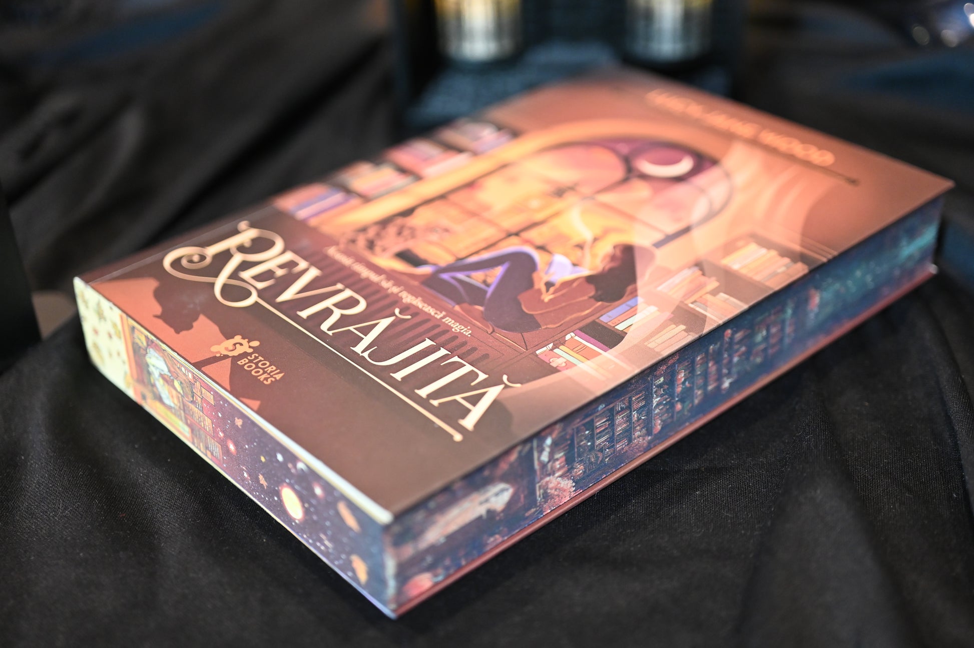 Cartea Revrăjita de Lucy Jane Wood, fotografiată în prim-plan pe un fundal întunecat. Coperta arată o tânără care citește lângă o fereastră luminată de lună, iar pe margini se văd detalii ilustrate cu rafturi de cărți și motive cosmice, în nuanțe de mov și cupru.