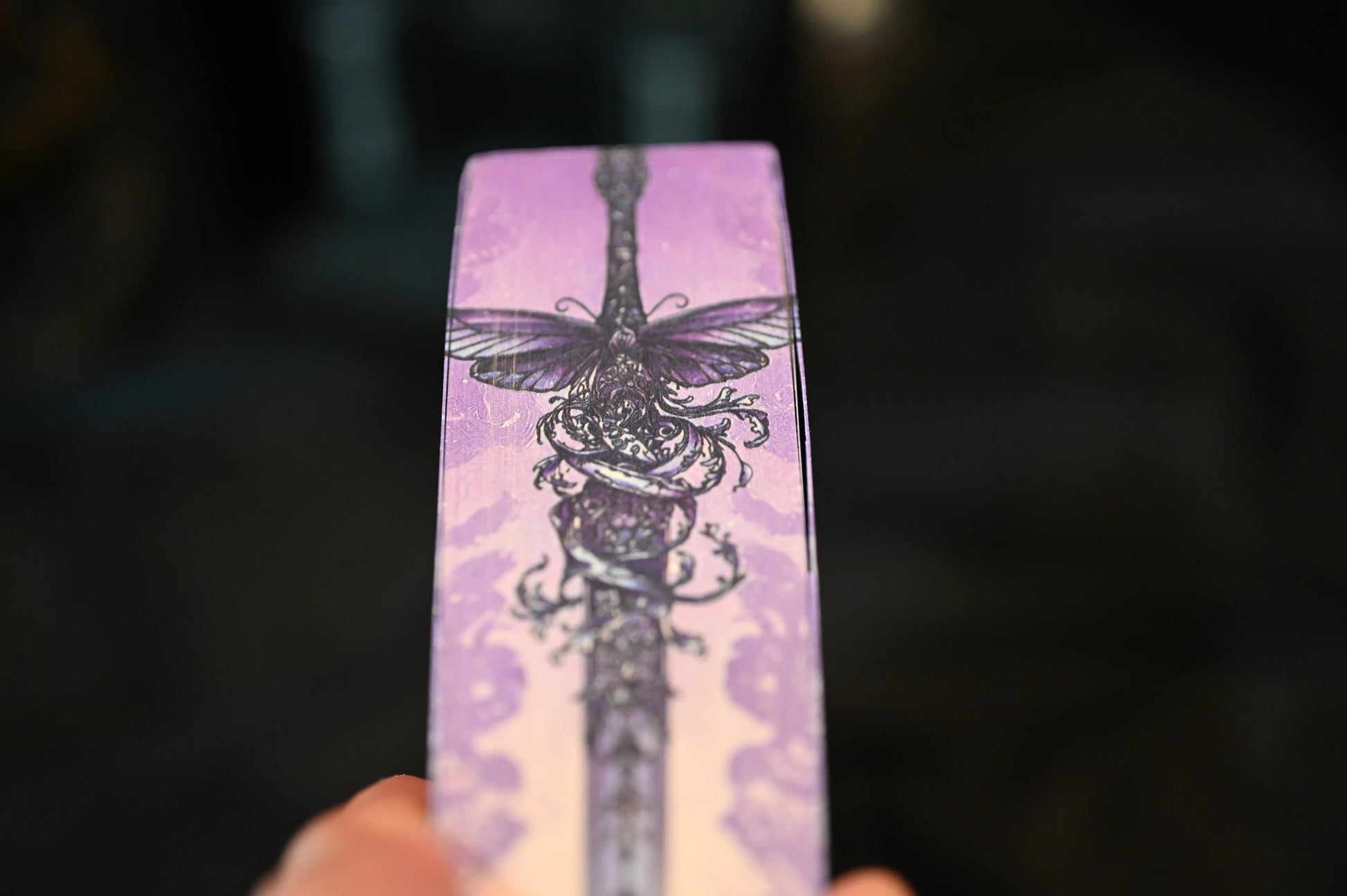 Detaliu de pe marginea superioară a cărții Cavalerul și Molia de Rachel Gillig. Ilustrația imprimată arată o sabie decorată cu o molie violet și ornamente florale pe fundal mov, într-un stil gotic-fantasy rafinat.