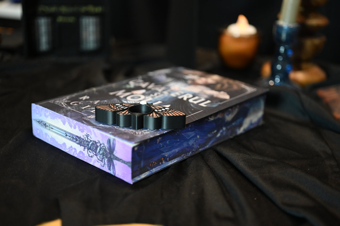 Detaliu de pe marginea superioară a cărții Cavalerul și Molia de Rachel Gillig. Ilustrația imprimată arată o sabie decorată cu o molie violet și ornamente florale pe fundal mov, într-un stil gotic-fantasy rafinat.