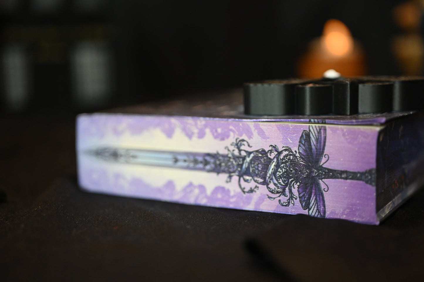 Detaliu de pe marginea superioară a cărții Cavalerul și Molia de Rachel Gillig. Ilustrația imprimată arată o sabie decorată cu o molie violet și ornamente florale pe fundal mov, într-un stil gotic-fantasy rafinat.