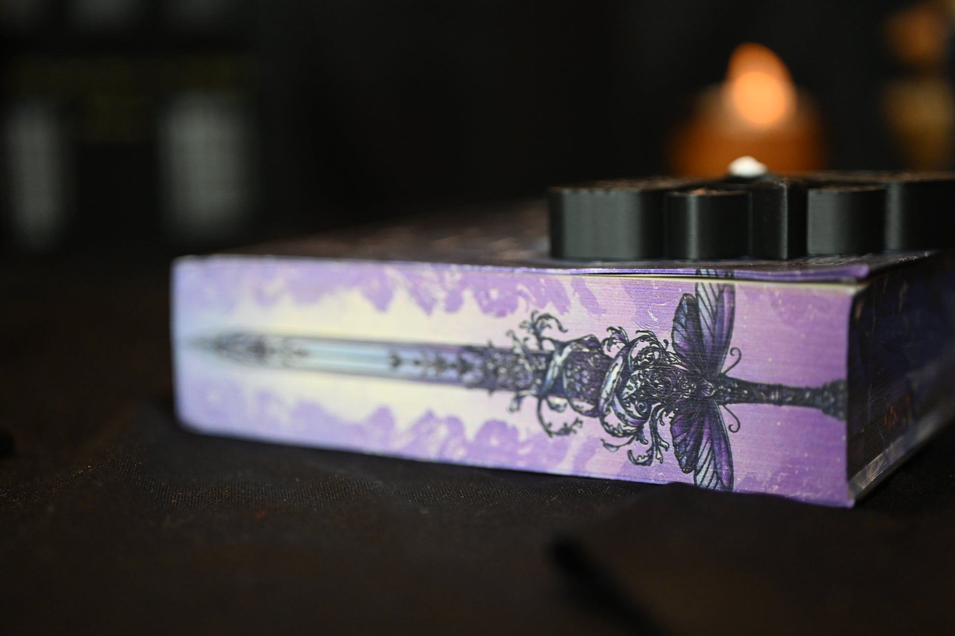 Detaliu de pe marginea superioară a cărții Cavalerul și Molia de Rachel Gillig. Ilustrația imprimată arată o sabie decorată cu o molie violet și ornamente florale pe fundal mov, într-un stil gotic-fantasy rafinat.