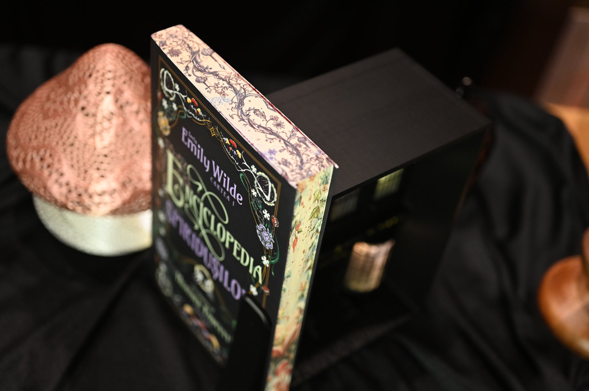 Vedere de sus a cărții Enciclopedia spiridușilor de Heather Fawcett, unde se observă detaliat designul imprimat pe margini: ramuri, frunze și flori desenate în linii fine negre și albastre, pe un fundal crem vintage.
