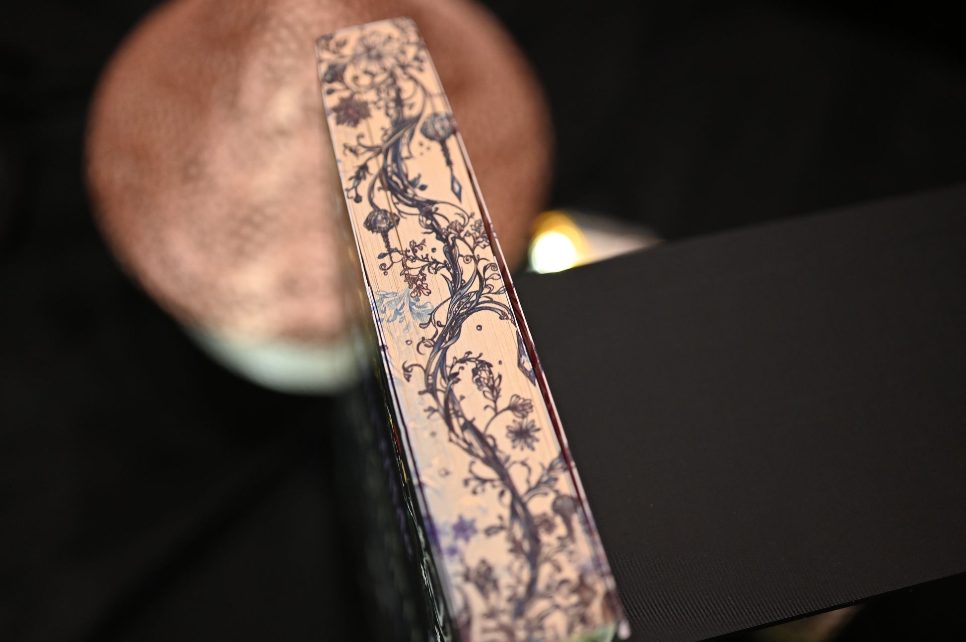 Vedere de sus a cărții Enciclopedia spiridușilor de Heather Fawcett, unde se observă detaliat designul imprimat pe margini: ramuri, frunze și flori desenate în linii fine negre și albastre, pe un fundal crem vintage.