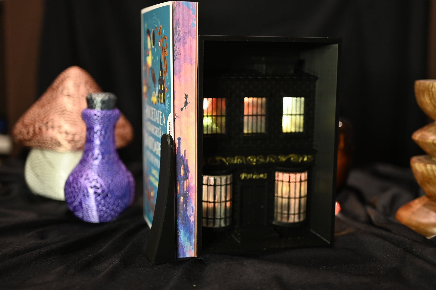 Cartea Societatea secretă a vrăjitoarelor de Sangu Mandanna, ediție specială cu margini ilustrate (printed edges). Marginea laterală a paginilor prezintă o casă iluminată sub cer roz-auriu, cu silueta unei vrăjitoare zburând pe mătură, în stil ilustrat magic.