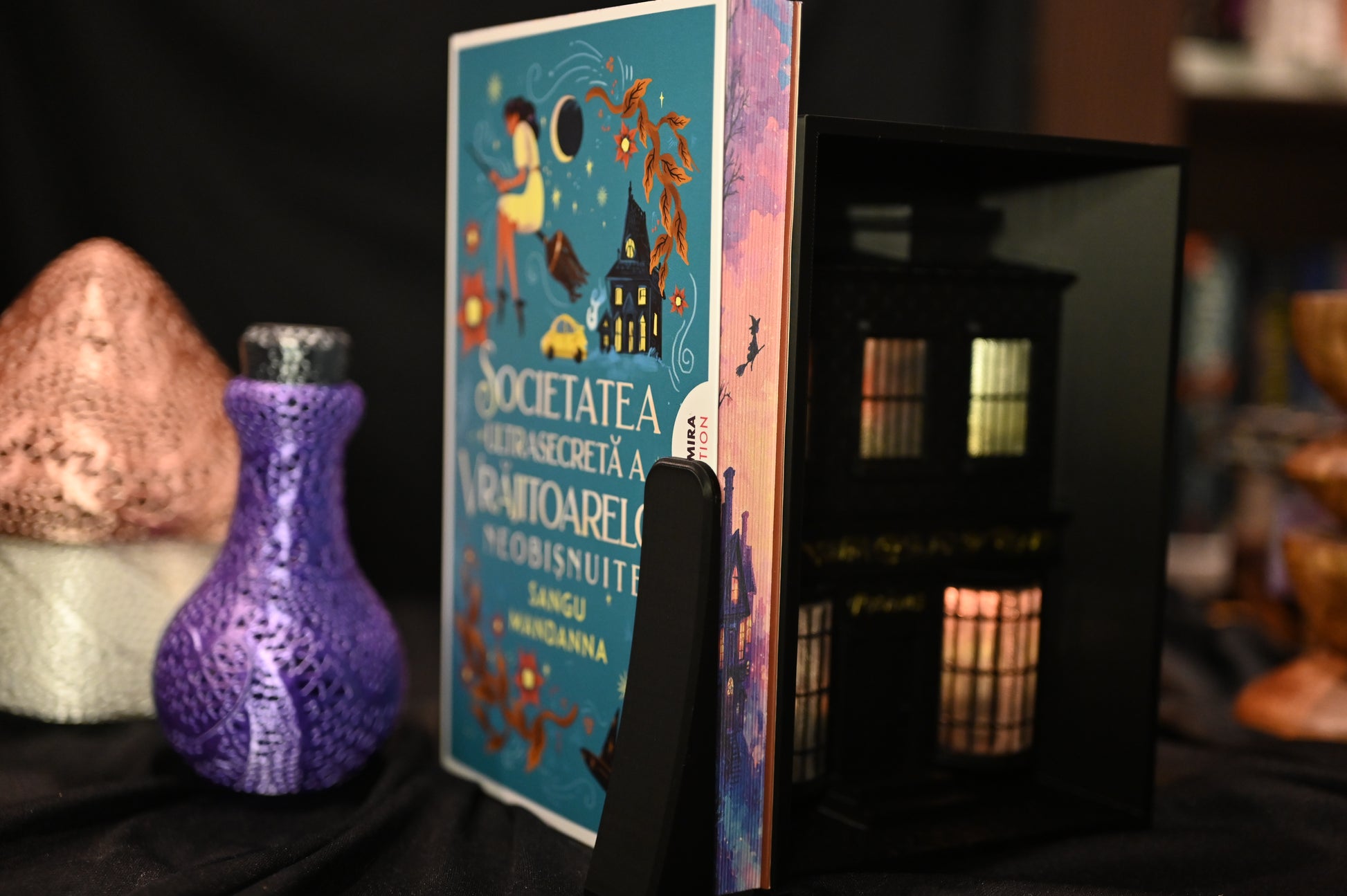 Cartea Societatea secretă a vrăjitoarelor de Sangu Mandanna, ediție specială cu margini ilustrate (printed edges). Marginea laterală a paginilor prezintă o casă iluminată sub cer roz-auriu, cu silueta unei vrăjitoare zburând pe mătură, în stil ilustrat magic.