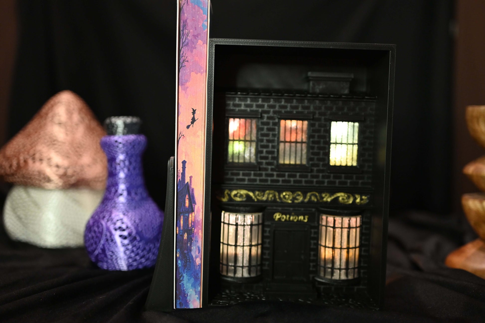 Cartea Societatea secretă a vrăjitoarelor de Sangu Mandanna, ediție specială cu margini ilustrate (printed edges). Marginea laterală a paginilor prezintă o casă iluminată sub cer roz-auriu, cu silueta unei vrăjitoare zburând pe mătură, în stil ilustrat magic.