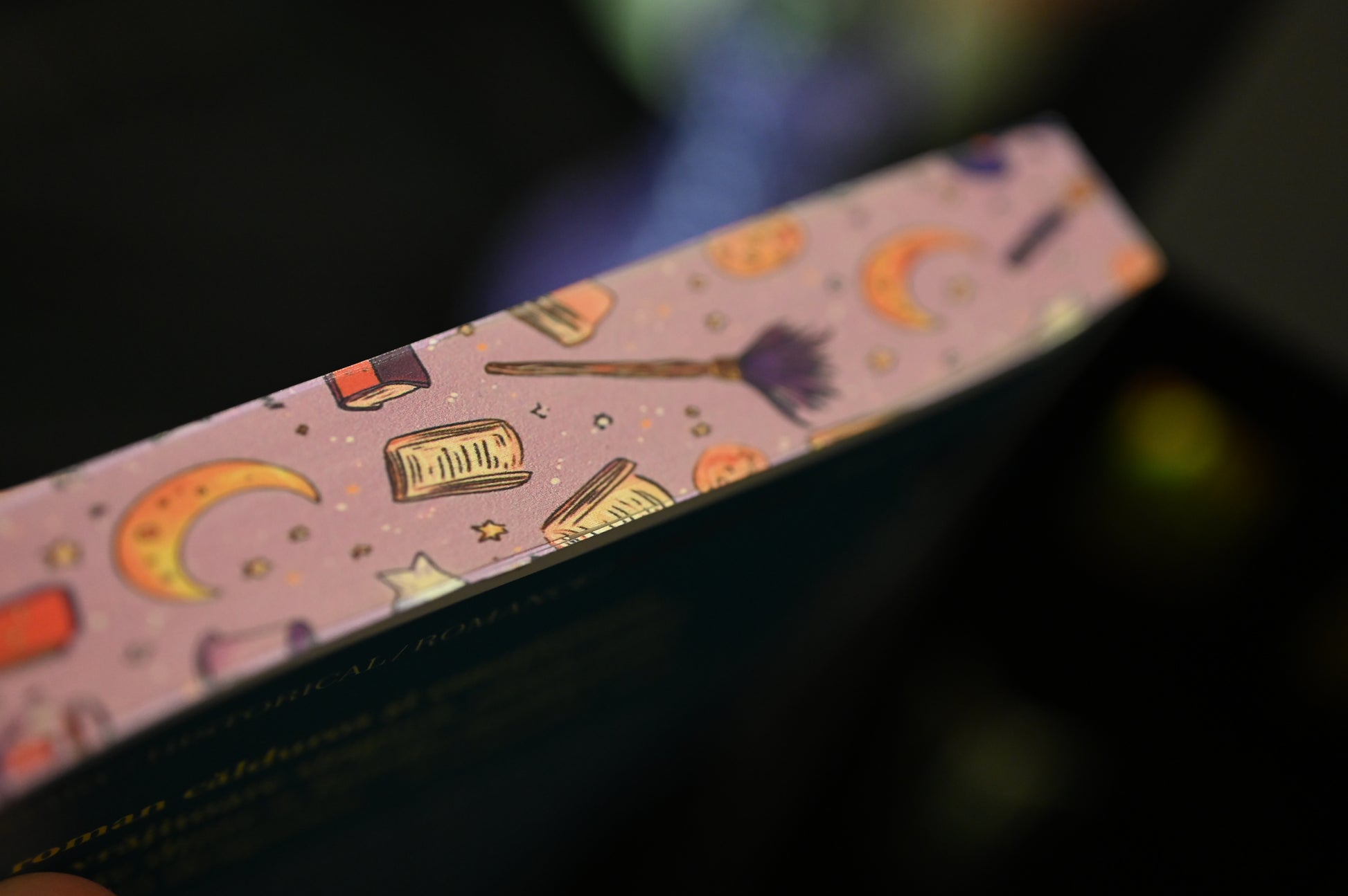 Vedere de sus a cărții Societatea secretă a vrăjitoarelor de Sangu Mandanna, unde marginea superioară este imprimată cu simboluri magice: cărți, stele, semilune și o mătură de vrăjitoare, pe un fundal violet pastelat.