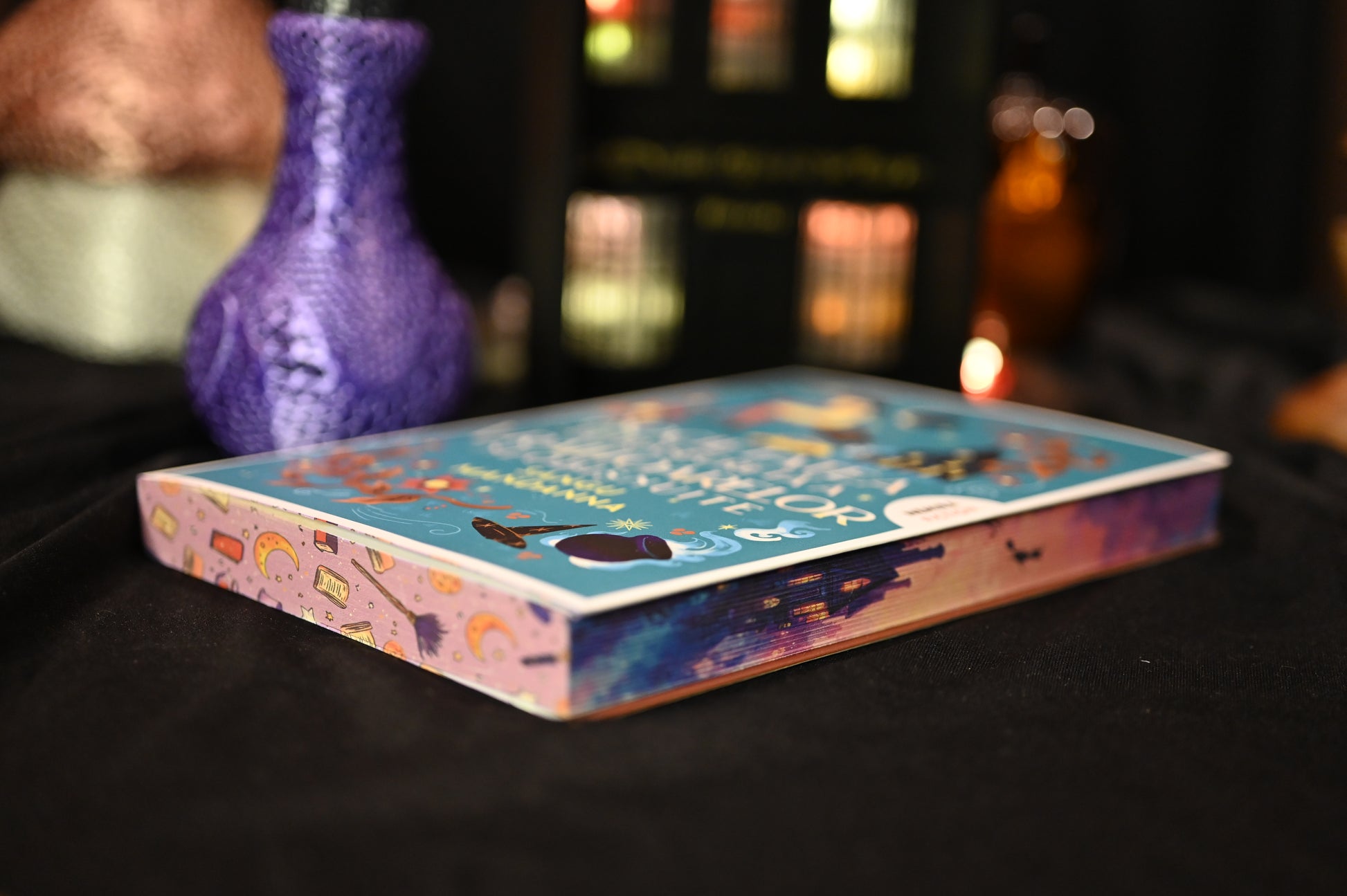 Cartea Societatea secretă a vrăjitoarelor de Sangu Mandanna, fotografiată în poziție orizontală. Coperta albastră vibrantă și marginea imprimată cu motive de magie, cărți și cer în nuanțe calde, accentuează tema fantasy și atmosfera cozy a ediției cu printed edges.