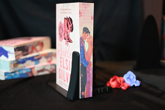 Cartea Reckless cu printed edges de Elsie Silver, ediție cu margini imprimate roz, înfățișează un cuplu apropiat: un bărbat brunet în cămașă cadrilată albastră și o femeie blondă în rochie roz, privind unul spre celălalt cu afecțiune. Marginea are o ilustrație cu cizme de