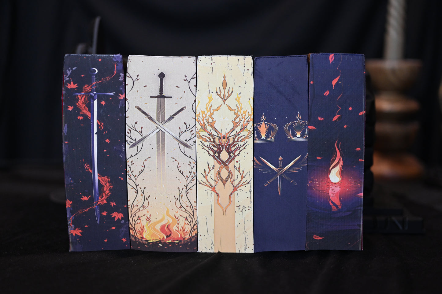 Set de cărți Din sânge și cenușă cu printed edges stilizate, prezentând designuri cu săbii încrucișate, coroane aurii, flăcări și ramuri decorative, în culori închise și tonuri calde.