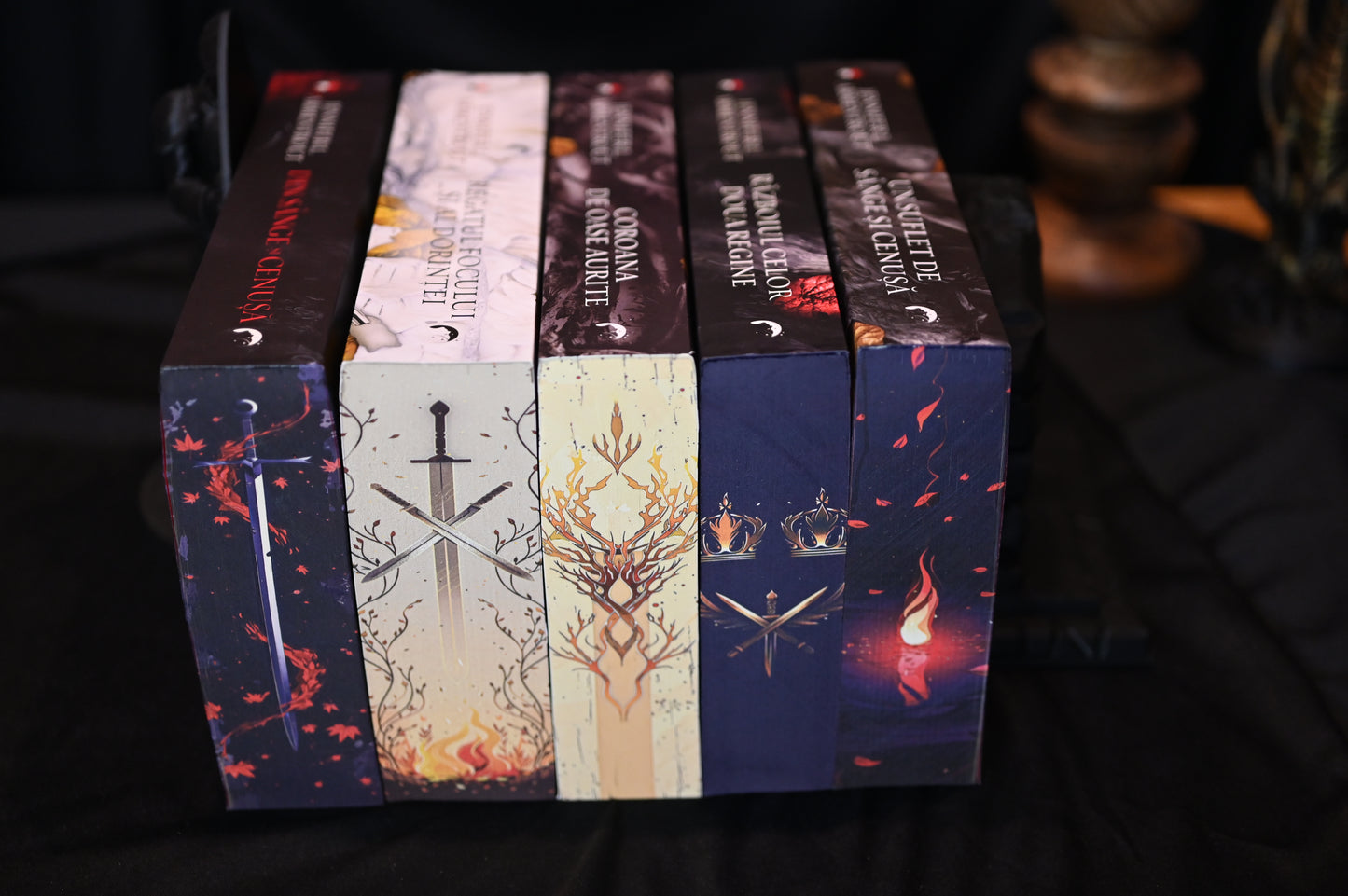 Set de cărți Din sânge și cenușă cu printed edges stilizate, prezentând designuri cu săbii încrucișate, coroane aurii, flăcări și ramuri decorative, în culori închise și tonuri calde.